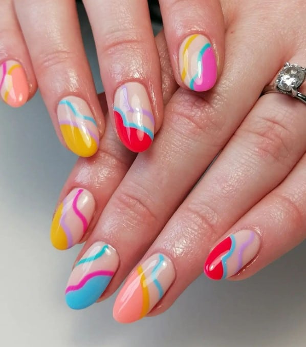 A closeup of a woman's oval-shaped nails with nude nail polish base that has whimsy sideway French tips that resemble fluffy clouds in red, blue, yellow, peach, and pink nail polish