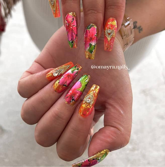 A woman's long coffin nails with gold foil smudges, pink hibiscus blooms, and brilliant studs and rhinestones