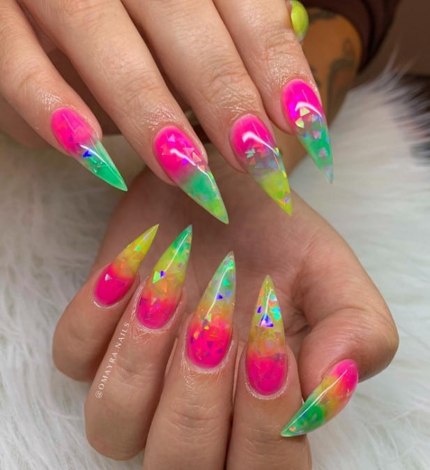 A woman's long stiletto nails with bright hues of pink, green, and yellow nail polish that has an encapsulated chromatic triangle glitter