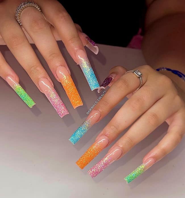 A woman's long square nails with nude nail polish base that has ombre colors, like green, purple, orange, pink, and blue in glitter nail tips