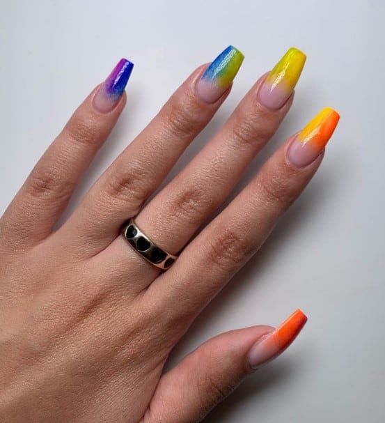 A woman's ballerina nails with nude nail polish base that has purple and blue, blue and yellow, and yellow and orange tips