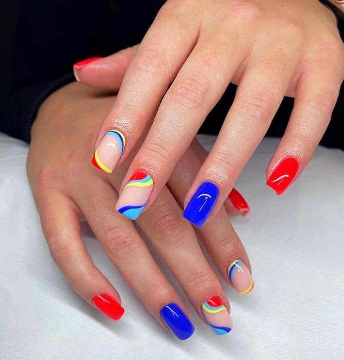 A closeup of a woman's long nails with nude, blue and red nail polish base that has swirls near the tips and the cuticles in multiple shades of blue, yellow, and red.
