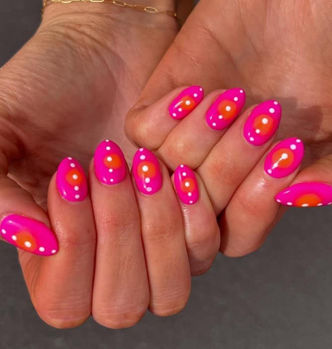 A closeup of a woman's almond shaped nails with a bright pink nail polish that has large orange circles at the center and vertical rows of white dots