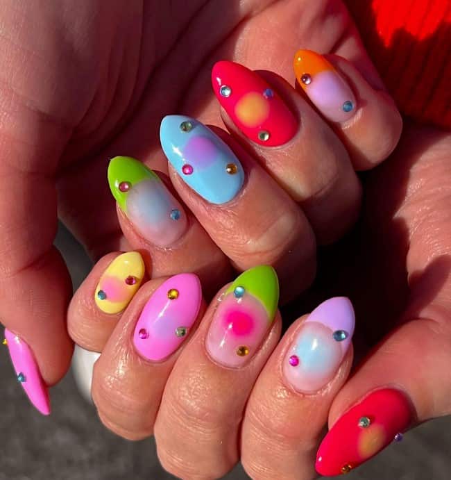 A closeup of a woman's long nails with vibrant color nail polish that has bright French tips in green, purple, and orange a soft-edged circle in the center and colorful gems