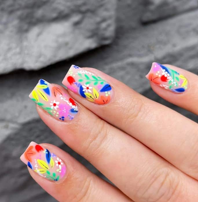 A closeup of a woman's square nails in matte nude fingernails with colorful garden by adorning each nail that has flowers and leaves in vibrant colors like yellow, red, orange, blue, and green