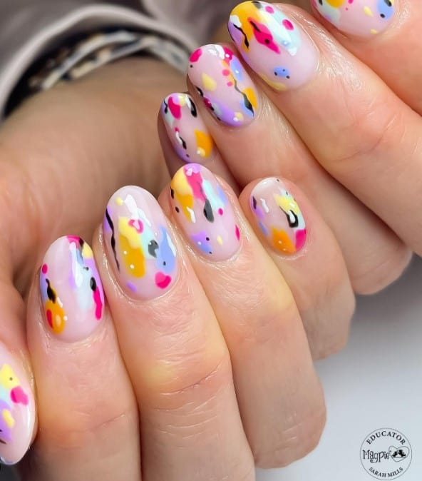 A closeup of a woman's short nails with nude nail polish that has splatters in black and assorted colors