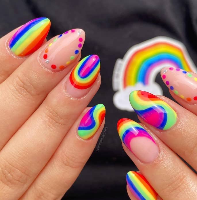 A closeup of a woman's almond shaped nails with rainbow inspired nail polish that has rainbow colors such as vertical lines, dotted outlines with gems, swirls, French tips, and rainbow art near the tips
