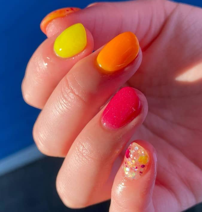 A closeup of a woman's short nails with yellow, orange, and red nail polish that has glitter on clear accent nails