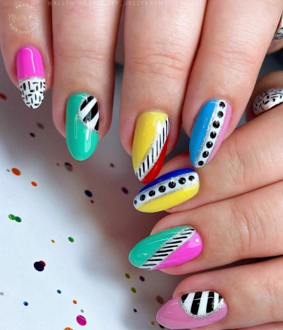 A closeup of a woman's long oval nails with bright colors like pink, green, yellow, red, and blue nail polish that has black and white nail art like basket weave, polka dots, and thick and thin diagonal lines in silver glitter