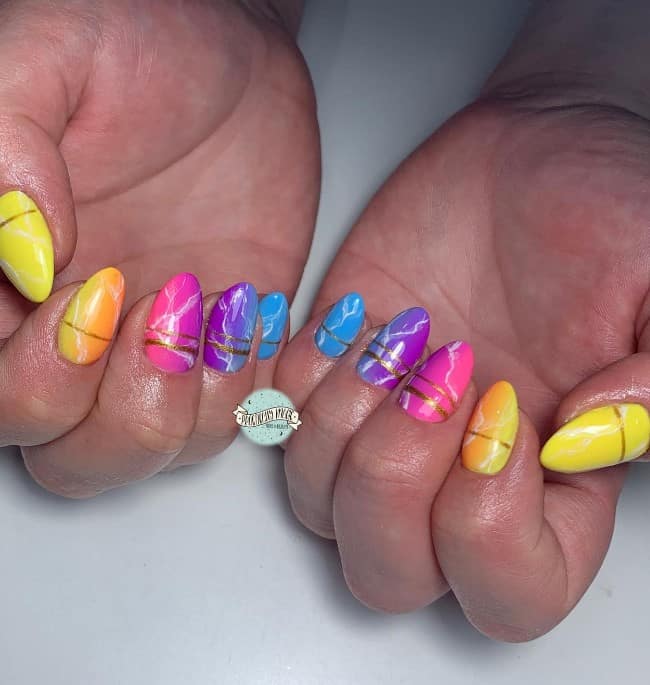 A closeup of a woman's mid-length nails with rainbow colors like yellow, orange, pink, indigo, and blue that transition vertically on the nails that has white marble effect and golden horizontal lines near the cuticles