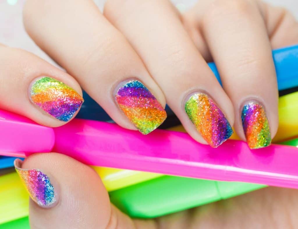 A closeup of a woman's short nails with rainbow inspired nail polish base that has glitter