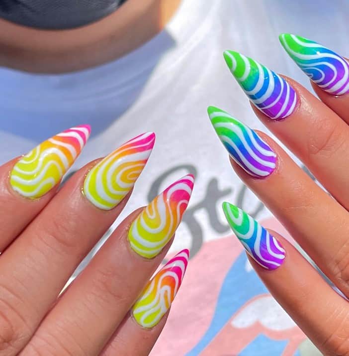 A closeup of a woman's stiletto nails with white nail polish base that has pink orange and yellow on the nails of one hand and green blue and purple on the other
