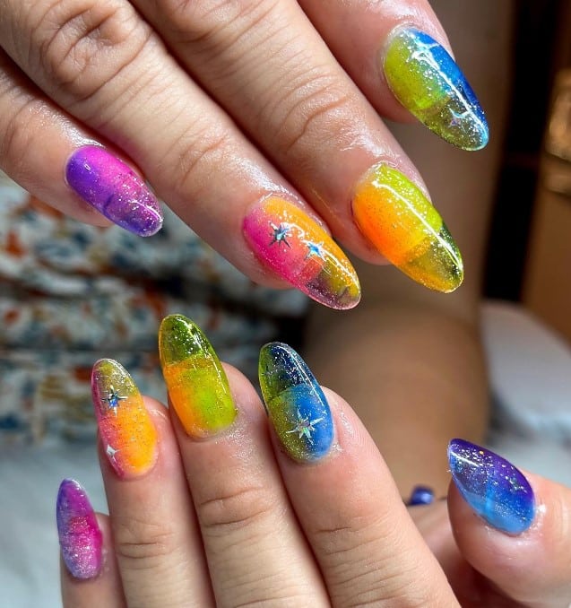 A closeup of a woman's long almond shaped nails with purple, orange, yellow, and blue nail polish base that has silver glitter and a silver star art