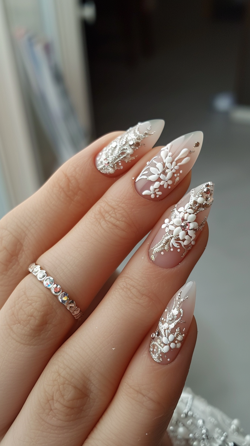 These stiletto nails feature a nude base with white 3D floral appliques and glittery silver leaf accents, complemented by small pearl and rhinestone details for a luxurious bridal look.