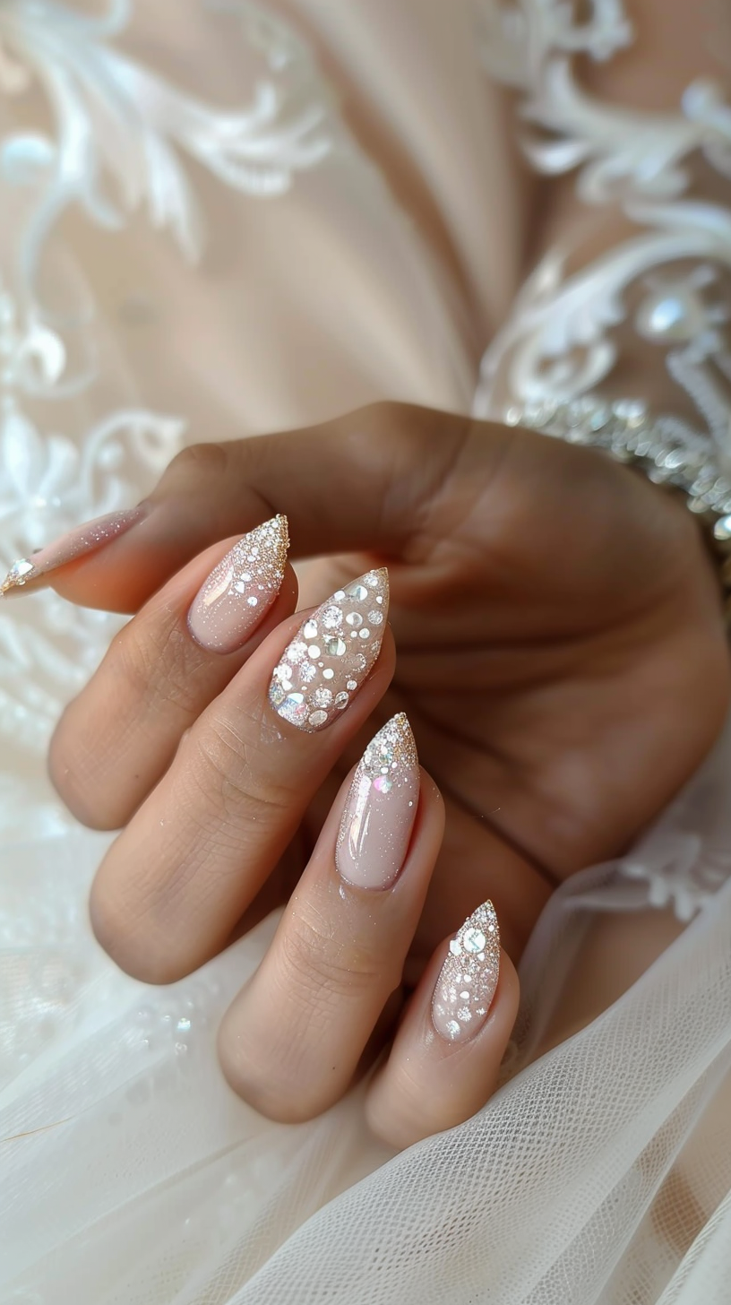 This bridal nail design exudes understated glamour with a nude base adorned with shimmering crystals and pearls. The nails feature a classic almond shape with a delicate pearl accent on one nail and clusters of sparkling gemstones scattered asymmetrically across the others.